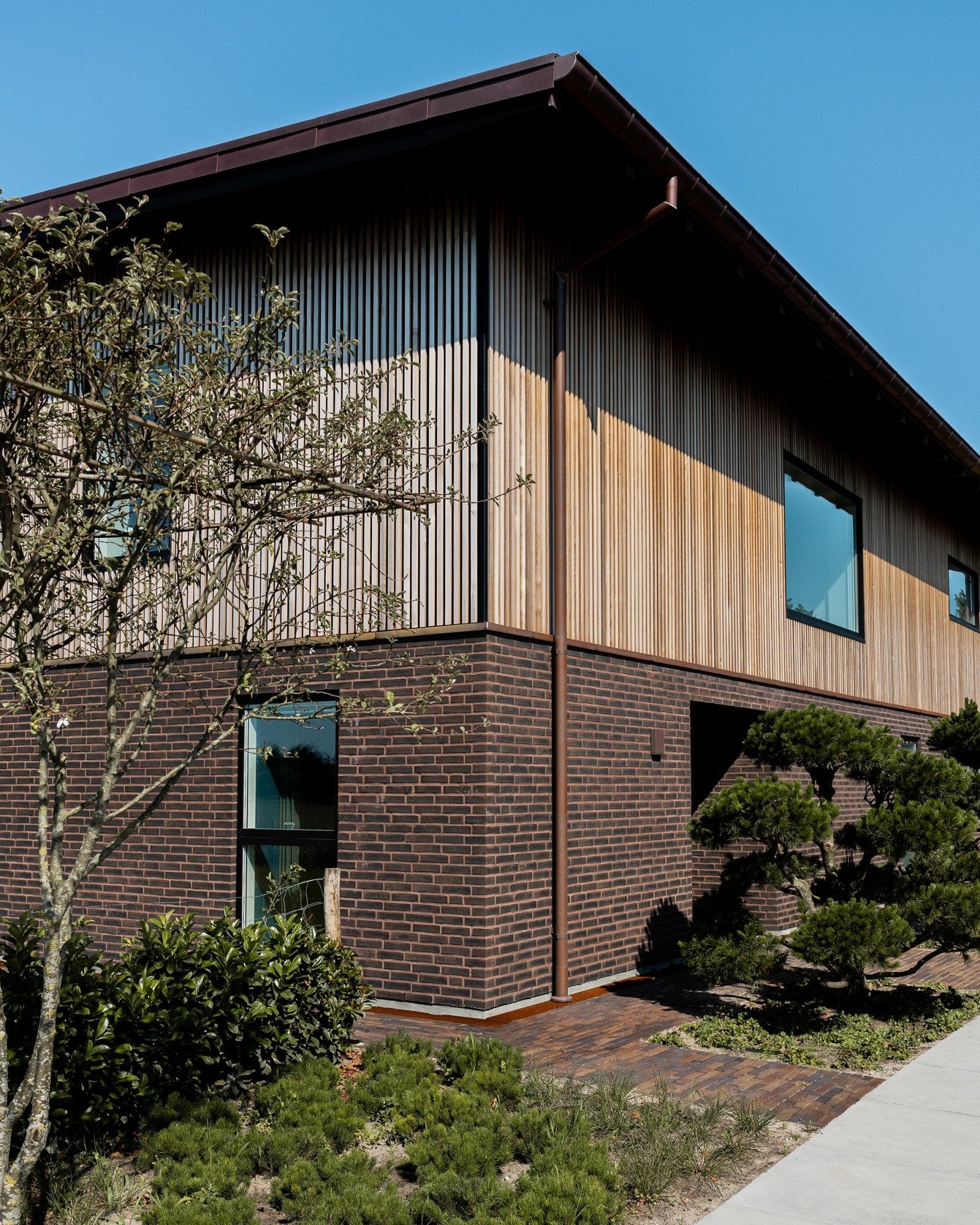 Maison à deux étages avec un rez-de-chaussée en brique et des panneaux de façade en bois vertical, entourée de verdure sous un ciel bleu dégagé.